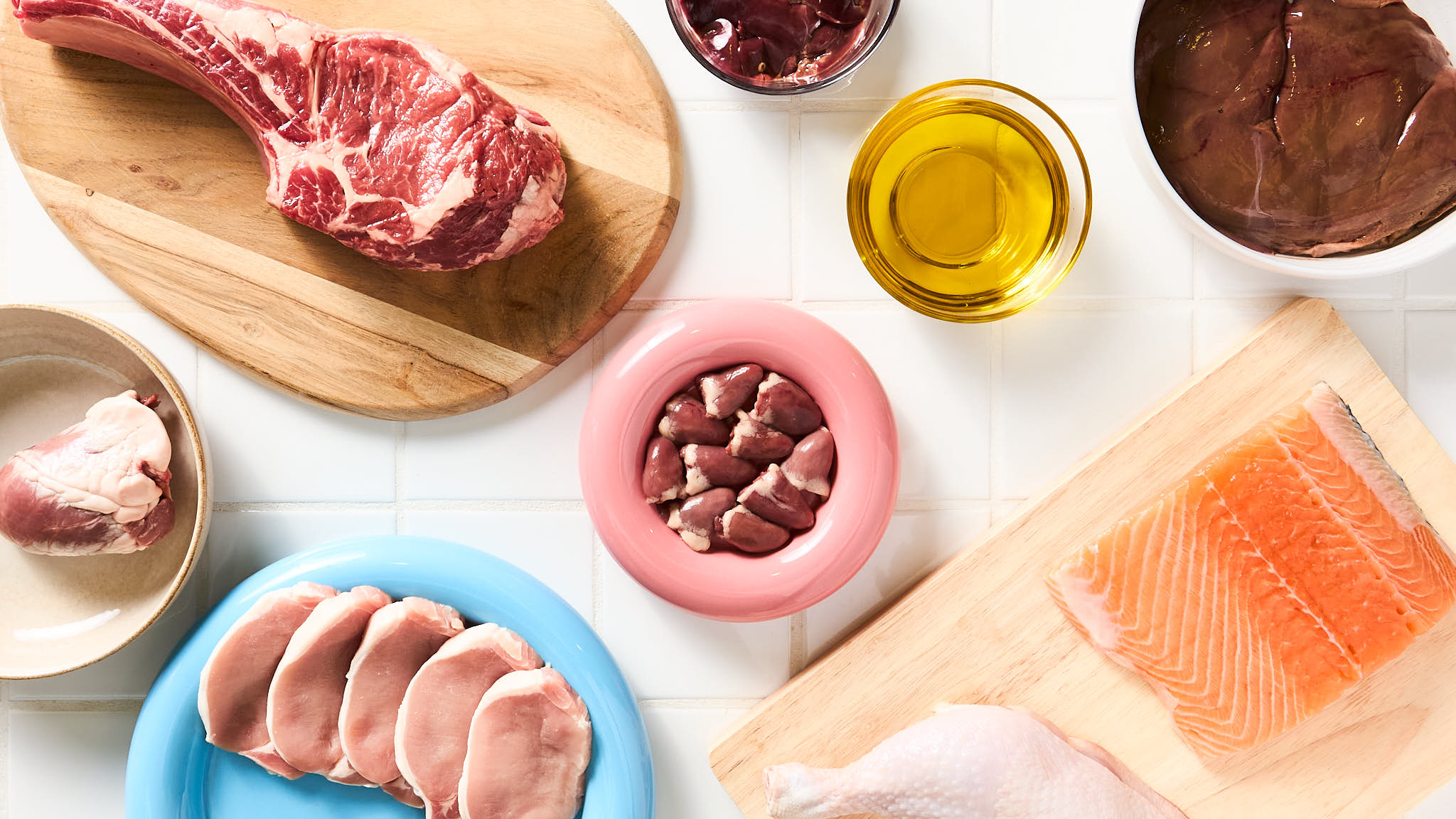 A top-down view of fresh, human-grade ingredients used in Pikko meals, including raw meats, salmon, chicken, organ meats, and oil on cutting boards. A top-down view of fresh, human-grade ingredients used in Pikko meals, including raw meats, salmon, chicken, organ meats, and oil on cutting boards.